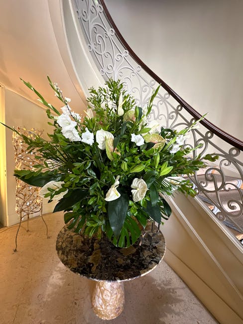 A large, fresh floral arrangement in a black ceramic vase featuring predominantly white flowers, including calla lilies, roses, and anthuriums, accented by lush green foliage such as broad leaves and 