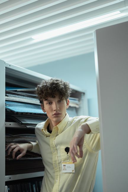 There is no image of flowers or floral arrangements provided; the image shows a young man with curly hair wearing a pale yellow shirt and a company badge, standing in an office environment near a shel