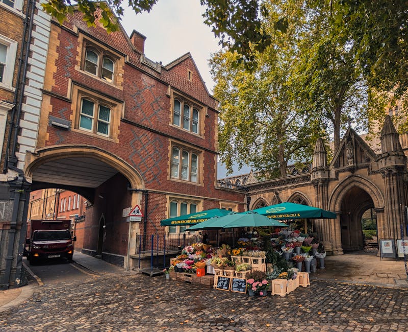 A flower stall situated outdoors near an ornate, historic stone archway and a red brick building with arched windows, set on a cobblestone street. The stall features a green canopy with branding, disp