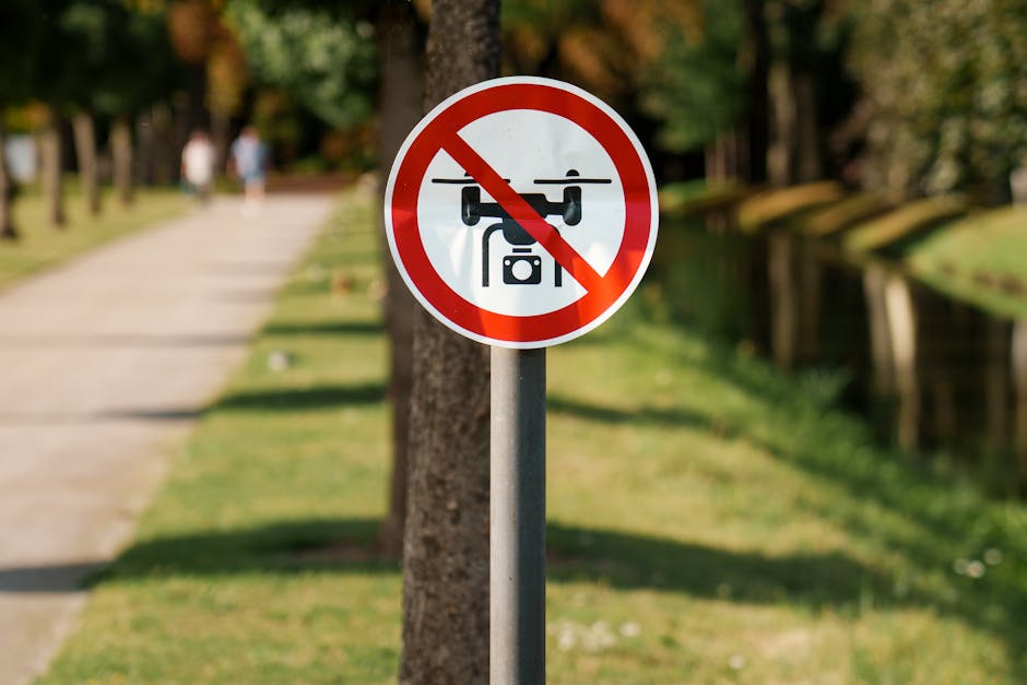 A circular traffic sign with a red border and a white background displaying a black drone with crossed lines indicating no drone flying is allowed. The sign is mounted on a metal pole situated along a